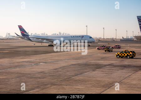 A LATAM Airlines Airbus A350, auf Leihbasis für Qatar Airways, Taxis zum Parkplatz am Flughafen Hamad, während Follow Me Autos auf dem Vorfeld geparkt sitzen Stockfoto