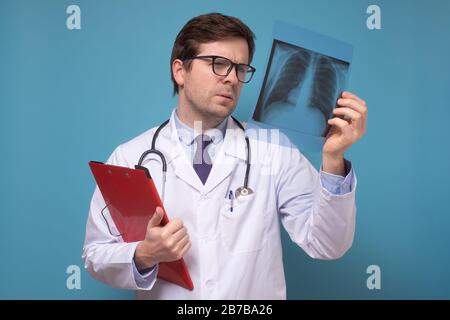 Der männliche Arzt, der das Röntgenbild der Lunge überprüft, konzentriert sich auf das Röntgenbild, das eine Lungenentzündung vermuten kann. Studio an blauer Wand gedreht. Stockfoto