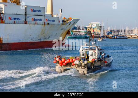 Ein kleines traditionelles Fischerboot wird von einem großen Containerschiff in der Nähe von Gosport im Portsmouth Harbour, Portsmouth, Hampshire, Südengland in den Schatten gestellt Stockfoto