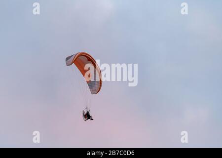 Mit Paragliding angetriebener Mann, der mit Fallschirm und Motor auf dem Rücken hoch in den Himmel fliegt. Der rosafarbene Himmel auf einem Hintergrund, Sonnenuntergang. Stockfoto