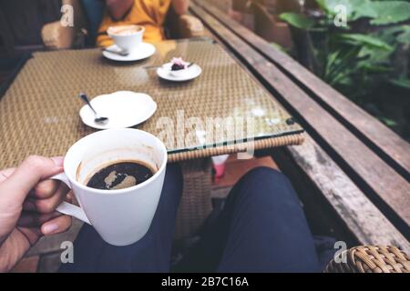 Nahaufnahme der Hand eines Mannes, der eine schwarze Kaffeetasse und eine Frau im Hintergrund im Café hält Stockfoto