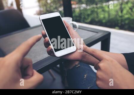 Verspottete Bilder von zwei Personen, die auf ein weißes Mobiltelefon mit leerem schwarzem Desktop-Bildschirm im Café blicken und zeigen Stockfoto