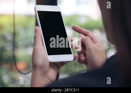 Verspottete Bilder von Frauen, die ein weißes Mobiltelefon mit leerem schwarzem Desktop-Bildschirm im Café halten und darauf zeigen Stockfoto