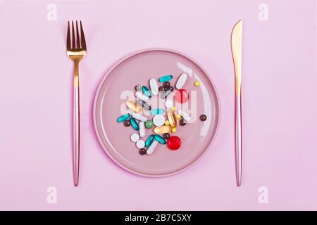 Bunte Kapseln und Pillen auf Platte mit Gabel und Messer auf rosa Hintergrund. Ernährung, Medizin, Gesundheitskonzept Stockfoto