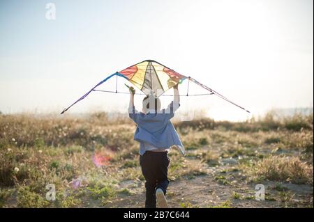 Aktiver kleiner Junge mit farbenfrohem Drachen, der in Richtung des hellen Sonnenuntergangs und des Seeufers läuft und zu fliegen versucht Stockfoto