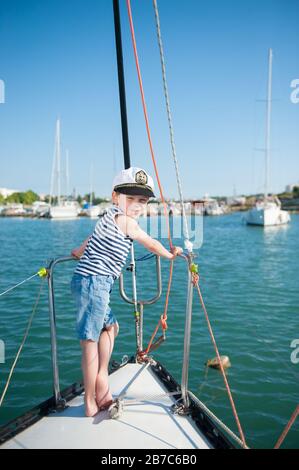 Fröhlicher süßer kleiner Junge in Kapitän weißer Hut gestreiftes Tanktop und Jeans-Shorts, die auf luxuriöser Meeresyacht im Hafen während der Sommerfahrt stehen Stockfoto