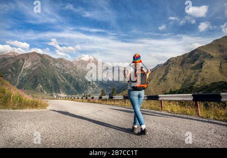 Touristenfrau mit Rucksack und Regenbogenhut, die in den Bergen in der Nähe des großen Almaty-Sees in Kasachstan die Straße hinuntergeht Stockfoto