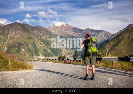 Tourist mit Rucksack und Regenbogenhut, der die Straße in den Bergen in der Nähe des Big Almaty Lake in Kasachstan hinunter geht Stockfoto