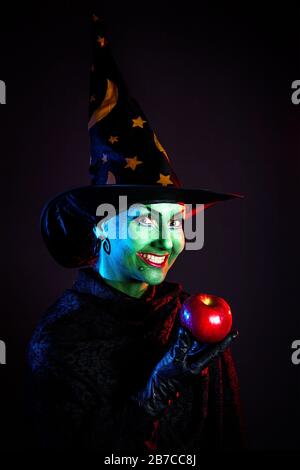 Hexe mit grüner Haut, die roten apfel hält und an Halloween auf dunklem Hintergrund lächelt Stockfoto