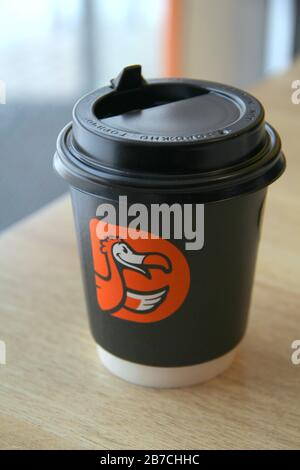 08-02-2020, Russland, Syktyvkar. Kaffeetasse aus schwarzem Papier mit schwarzem Kunststoffdeckel und orangefarbenem Logo der internationalen Pizzeria Kette. Dodo Pizza steht auf dem Tisch in der Nähe des Fensters im Restaurant. Stockfoto