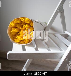 Auf einem Stuhl liegt ein in weißes Handwerkspapier gehüllter Blumenstrauß aus gelben Tulpen Stockfoto