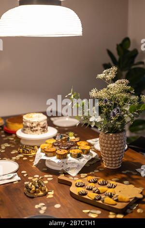 Dekorierter Geburtstagstisch mit verschiedenen Cupcakes, Muffins, Kuchen und Madeleinen mit Blumen und Konfetti. Stockfoto