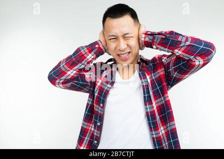 Gutaussehender asiate in einem karierten Hemd, das weit offen ist, bedeckt seine Ohren vor Lärm auf einem weißen Studiohintergrund Stockfoto