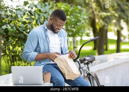 Mittagessen Im Park. Black Guy, Der Im Freien Mit Laptop Isst Und Ausruht Stockfoto