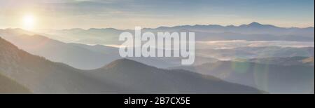 Panorama alpine Landschaft der Berggipfel und Täler, die im Morgengrauen von Nebel bedeckt sind. Frühlingslandschaften Berge und bunter Wald auf dem Hintergrund Sonne Stockfoto