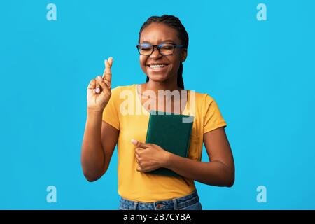 Student Girl Keeping Fingers Crossed Holding Books Over Blue Background Stockfoto