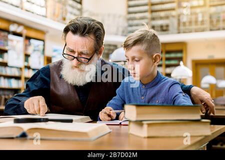 Der Senior belauscht den Mann, den Lehrer oder den Großvater, der das Buch laut vorliest, vor seinem Enkel oder Schüler, der ihm aufmerksam zuhört und Notizen macht. Gr Stockfoto