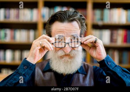 Gutaussehender lächelnder älterer bärtiger Mann, der seine Brillen berührt, in der Bibliothek oder im Buchladen über dem verschwommenen Hintergrund von Bücherregalen steht. Nahaufnahme p Stockfoto