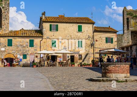 Ein Brunnen inmitten der Piazza Roma, Monteriggioni, Toskana, Italien, umgeben von historischen Gebäuden, einigen Wohnhäusern, Restaurants und Gelaterien Stockfoto