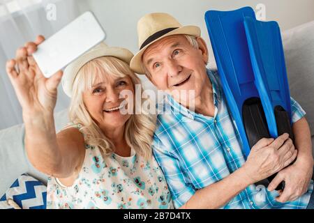 Leitender Mann und Frau sitzen im Reisebüro auf dem Sofa und tragen Strandhüte Ehemann, der Flipper hält, während die Frau das Smartphone bei selfie hält Stockfoto