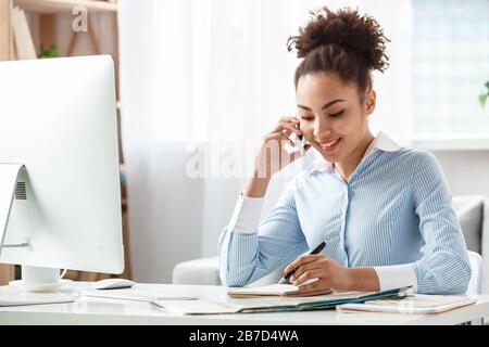 Afrikanischer Touragent mit weiblicher Abstammung sitzt am Tisch im Büro und spricht mit dem Kunden, der sich Notizen über Clien nimmt, muss fröhlich lächeln Stockfoto
