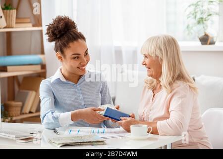 Afrikanischer Absteiger, weiblicher Reisevermittler im Büro, der Zahlung und Reisepass von einer älteren Frau abnimmt, die fröhlich lächelt Stockfoto
