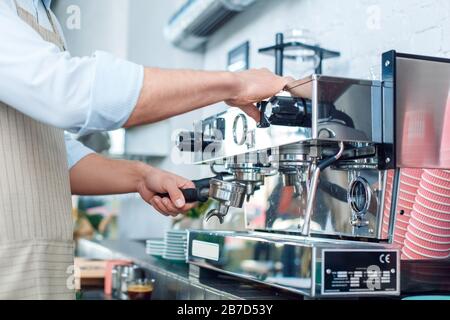 Junger, gutaussehender Barista, der in der Café-Bar steht und den Portafilter in die Kaffeemaschine einfügt, damit der Espresso aus nächster Nähe zubereitet wird Stockfoto