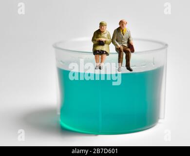 Miniatur-Figurin-Rentner sitzen auf einem Plastikbecher für flüssige Medikamente vor weißem Hintergrund. Stockfoto