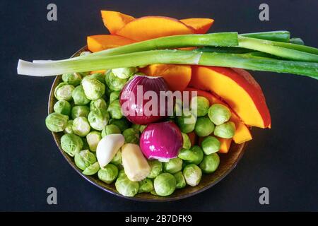 Platte mit einem bunten Sortiment an frischem Gemüse: Rosenkohl, rote Zwiebeln, Frühlingszwiebeln, Kürbis, Knoblauch und Ingwer. Schwarzer Hintergrund. Stockfoto