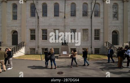 Massachusetts, USA. März 2020. Besucher fotografieren mit der Skulptur des Gründers der Harvard University John Harvard im Harvard Yard, Zentrum des Harvard Campus, das Gesicht von Harvard wurde mit einer Gesichtsmaske bedeckt. Als Reaktion auf Coronavirus Bedenken gab die Harvard University bekannt, dass ab 23. März 2020 alle Klassen remote oder online sein werden. Studenten, die in Studentenwohnheimen leben, wurden gebeten, vor dem 22. März 2020 zu gehen. Stockfoto