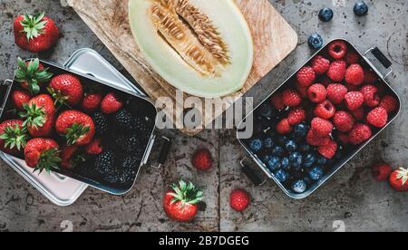 Gesundes veganes Sommeressen. Flat-lay von frischen, saisonalen Erdbeeren, Himbeeren, Blaubeeren, Brombeeren in Metalllunchboxen und Melone über rauem g Stockfoto