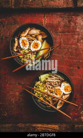 Flat-lay von traditionellen japanischen Ramen-Schüsseln mit Hühnerfleisch und Shiitake-Pilzen über dunkelrot beschädigten Holztischhintergrund, Draufsicht. Japanes Stockfoto