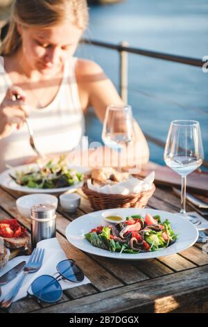 Schöne junge blonde Frau, die Salat mit Meeresfrüchten, Bruschetta und Wein im Sommercafé im Freien in Italien mit malerischem Blick auf den Hintergrou isst Stockfoto