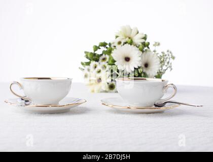 Frische weiße Gänseblümchen und grüne Blätter schmücken den Tisch für Teezeit mit antiken Tassen und Untertöpfen auf weißem Hintergrund mit Raum Stockfoto