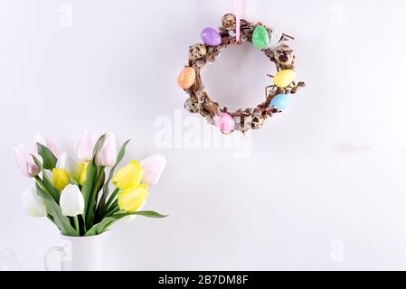 Osterkarte mit handgefertigtem Kranz aus Weidenzweigen und bemalten bunten Eiern, Tulpen-Blumen in Vase gegen hellgraue Wand. Stockfoto