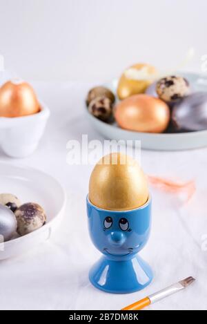 Handgemaltes goldenes Ei in einem blauen Becher als Gesicht auf einem textilen hellgrauen Hintergrund. Stockfoto