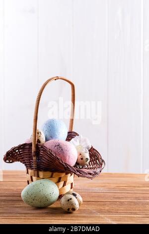 Festlicher Oster-Korbkorb mit handgemalten Hühnchen- und Wachteleiern auf einem Holztisch. Stockfoto