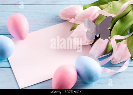 Frohe Ostern festliche Karte aus bemalten Eiern, Papierblatt und handgefertigten Schmetterlingen auf hellblauem Holzhintergrund. Stockfoto