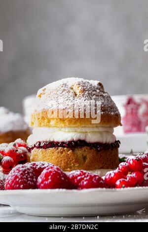 Victoria-Schwamm-Kuchen mit Himbeerbmarmelade und Schlagsahne, großzügig mit Puderzucker bestreut, serviert mit Früchten. Stockfoto