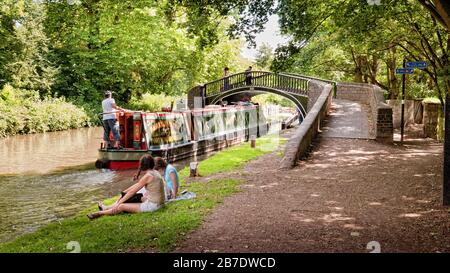 Schmalboot fährt unter der Isis-Fußgängerbrücke in die Isis-Schleuse, während Picknickfans den Oxford Canal (South), Oxford, England, Großbritannien, beobachten Stockfoto