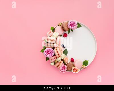 Schöne Komposition mit französischen Makronen, Blumen und Beeren auf Handwerksplatte über rosafarbenem Hintergrund. Wunderschönes, kreatives Layout mit Pastellmakaronen oder m Stockfoto