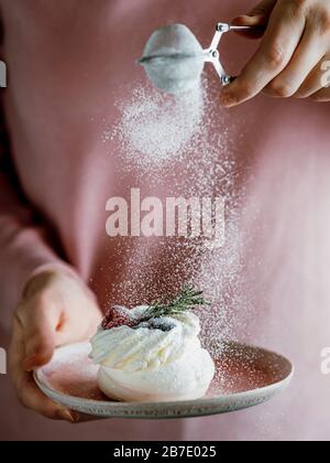 Frau in rosafarbener Bluse hält mit einer Hand rosa Platte mit Mini-Pavlova-Kuchen dekoriert frische Beeren und Rosmarin, und bestreut mit ihrer anderen Hand ici Stockfoto