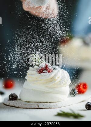 Frau bestreut Puderzucker durch ein Sieb auf einem kleinen Pavlova-Kuchen, der frische Beeren und Rosmarin verziert ist. Vertikale Zusammensetzung. Stockfoto