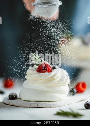 Frau bestreut Puderzucker durch ein Sieb auf einem kleinen Pavlova-Kuchen, der frische Beeren und Rosmarin verziert ist. Vertikale Zusammensetzung. Stockfoto