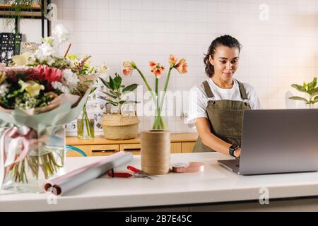 Frau trägt Schürze, die in ihrem Blumenladen arbeitet. Junge Geschäftsfrau tippt in geschlossenen Räumen auf Laptop. Stockfoto
