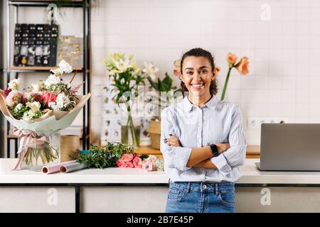 Selbstbewusste Floristin, die mit gekreuzten Armen steht und die Kamera betrachtet. Fröhlicher Blumenladen, der an der Theke steht. Stockfoto