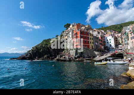 RIOMAGGIORE, ITALIEN - 15. August 2019: Blick auf Riomaggiore und das Meer mit Menschen, die an einem sonnigen Tag auf der Cinque Terre schwimmen Stockfoto