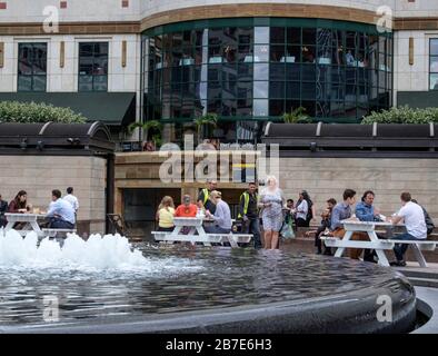 Die Büroangestellten in London essen an einem warmen Julitag Mittagessen an externen Tischen neben dem Brunnen am Cabot Square Canary Wharf, London. Stockfoto