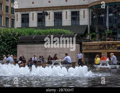 Die Büroangestellten in London essen an einem warmen Julitag Mittagessen an externen Tischen neben dem Brunnen am Cabot Square Canary Wharf, London. Stockfoto