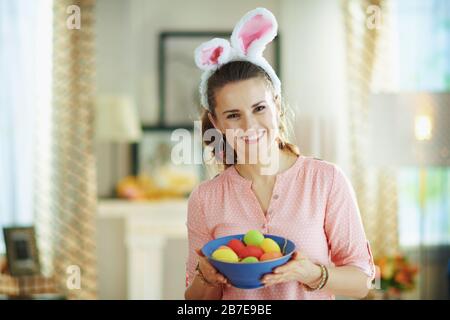 Porträt einer lächelnden eleganten 40 Jahre alten Hausfrau in einer rosafarbenen Bluse und osterhirnen, die einen blauen Teller mit bunten ostereiern in der Moderne halten Stockfoto